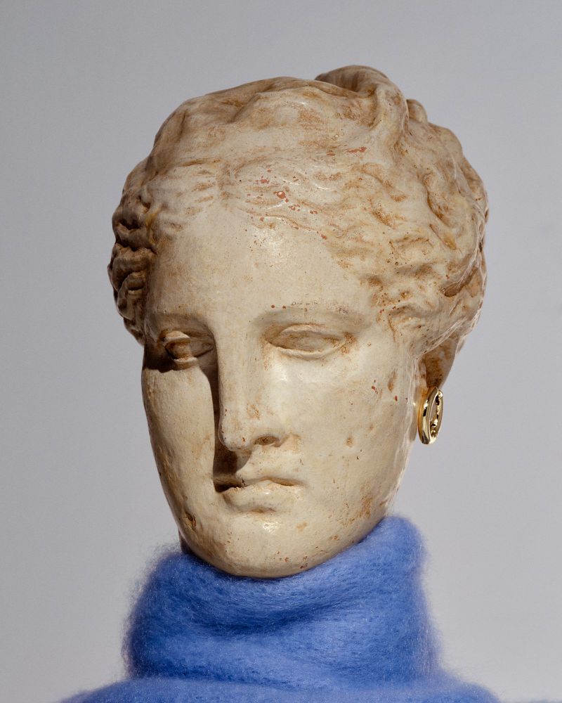 Bust sculpture of a female figure with a blue knit scarf, neutral background, stone texture, and gold earrings. Art and sculpture.
