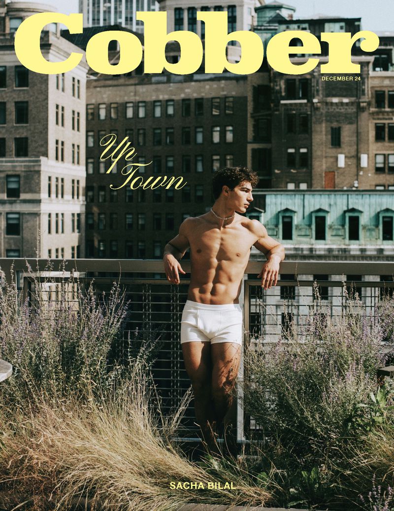 Magazine cover of a shirtless male model standing on a rooftop with city buildings in the background and lavender in the foreground.