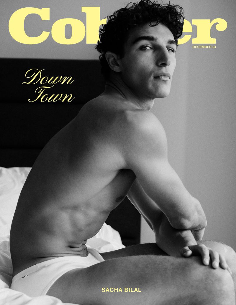 Black and white portrait of shirtless male model, SACHA BILAL, seated on a bed in underwear. Magazine cover with yellow text.