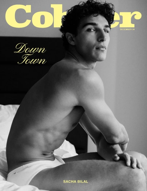 Black and white portrait of shirtless male model, SACHA BILAL, seated on a bed in underwear. Magazine cover with yellow text.
