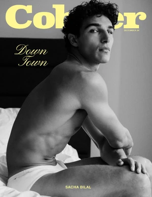 Shirtless male model Sacha Bilal seated on a white bed in a black and white magazine cover portrait