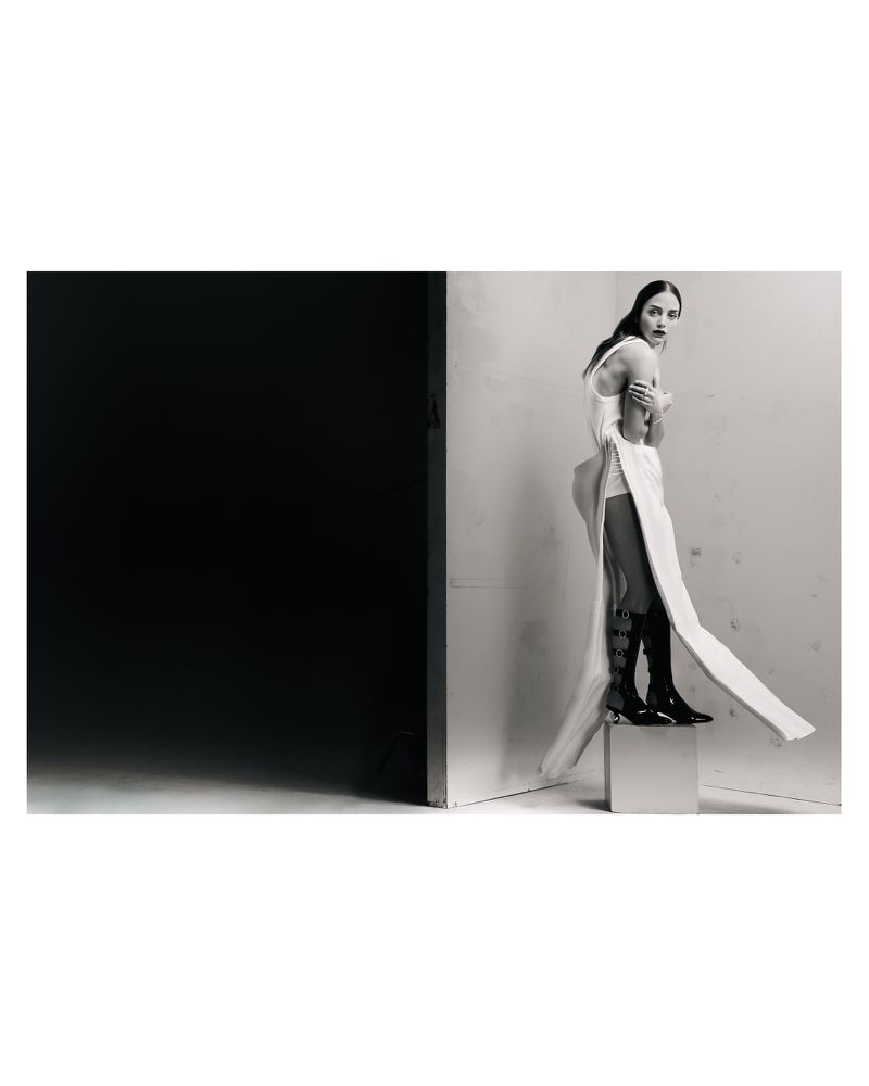 A woman in a white dress and high-heeled boots poses on a white cube in front of a contrasting black and white backdrop.