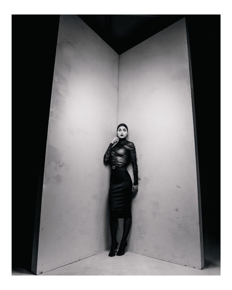 Monochrome portrait of a woman in a long-sleeved top and pencil skirt, posing against a corner in a dimly lit studio.