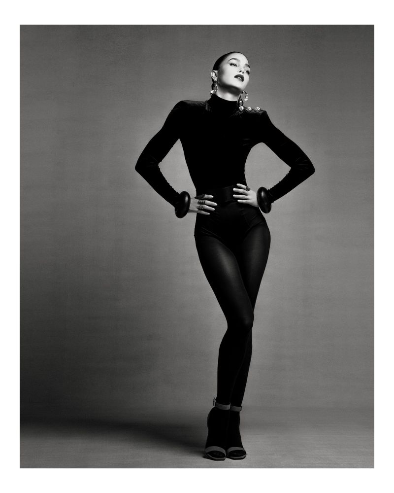 Monochrome studio shot of a female model in a black bodysuit, posing with hands on her hips and looking up.