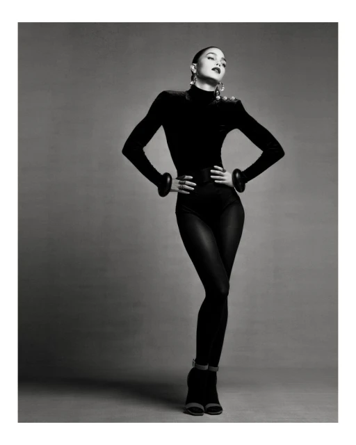 Monochrome studio shot of a female model in a black bodysuit, posing with hands on her hips and looking up.