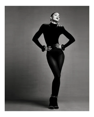 Monochrome studio shot of a female model in a black bodysuit, posing with hands on her hips and looking up.