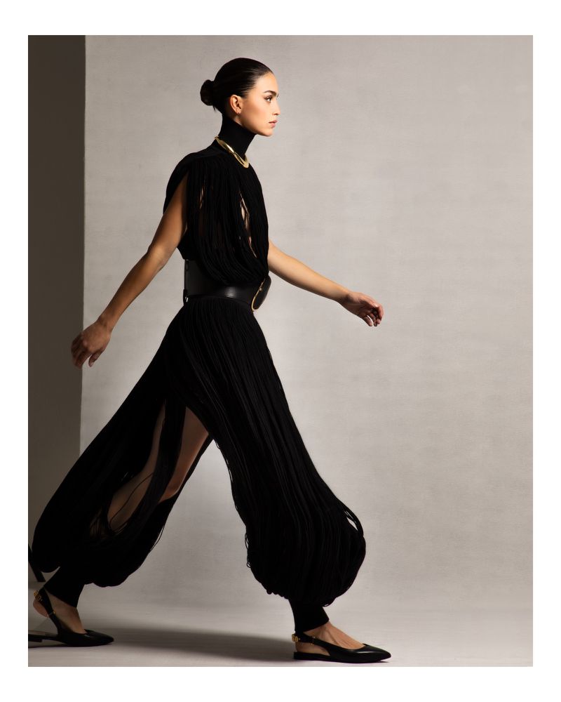 Fashionable woman in a black fringe dress walking in a studio. Model with dark hair and elegant attire striking a pose against a neutral backdrop.