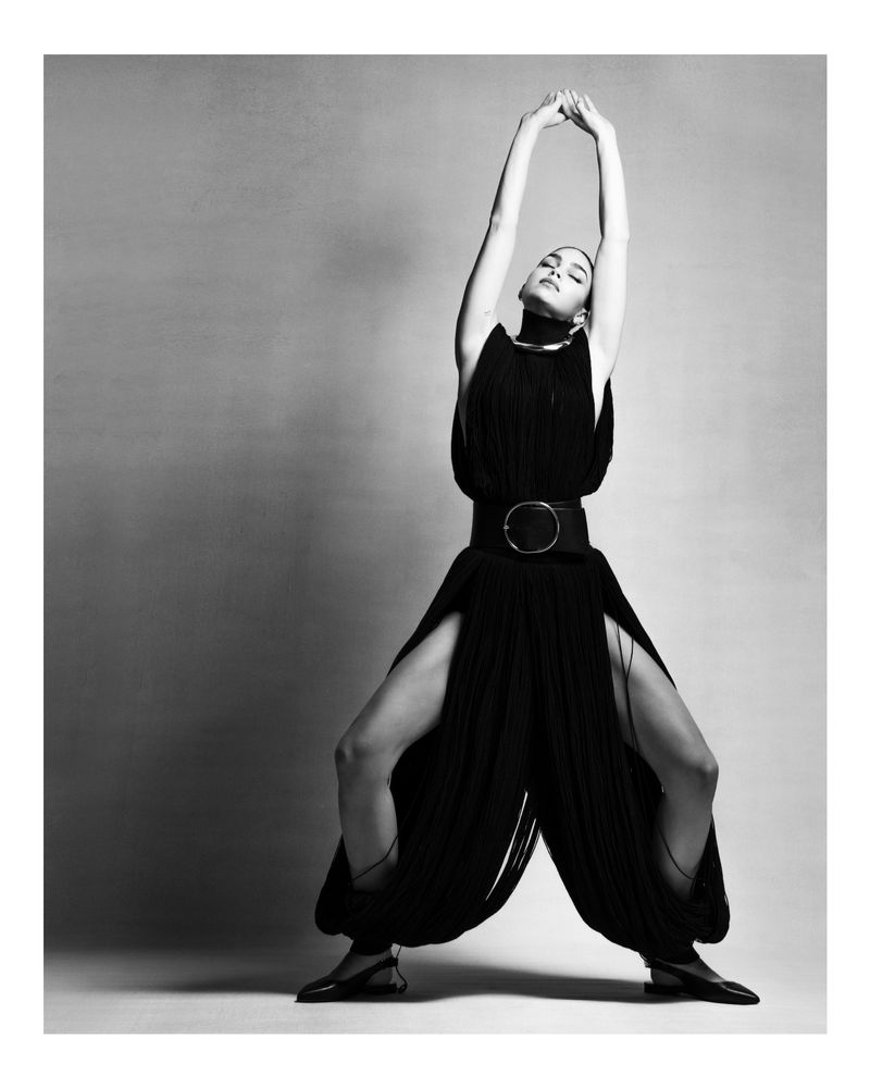 Monochrome studio portrait of an adult female with arms raised, wearing a black dress with a wide belt and black flats in a wide stance.