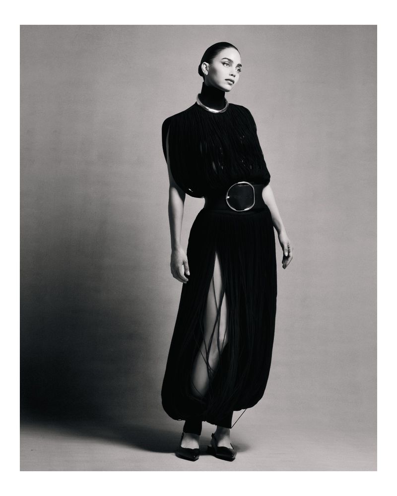 A woman in a black dress with a silver choker and belt poses in front of a neutral background in a studio fashion shot.