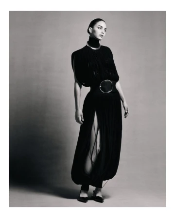 A woman in a black dress with a silver choker and belt poses in front of a neutral background in a studio fashion shot.