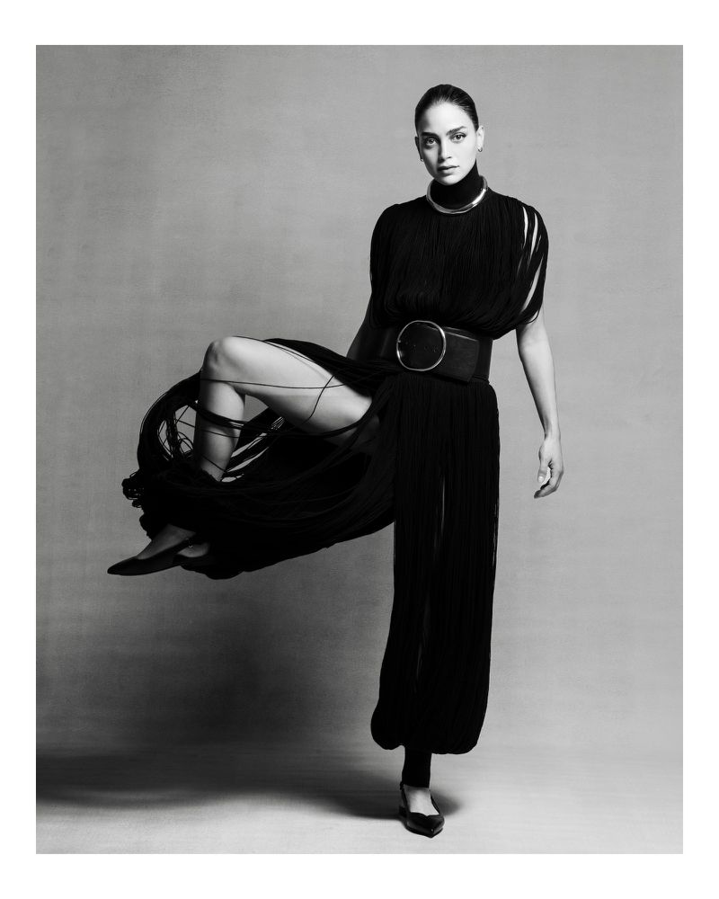 Monochrome studio portrait of a woman in an elegant black outfit with a high collar and wide belt, striking a dynamic pose.