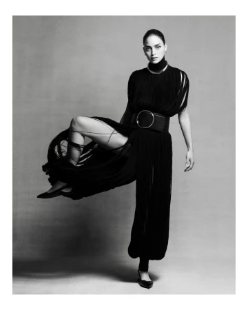 Monochrome studio portrait of a woman in an elegant black outfit with a high collar and wide belt, striking a dynamic pose.