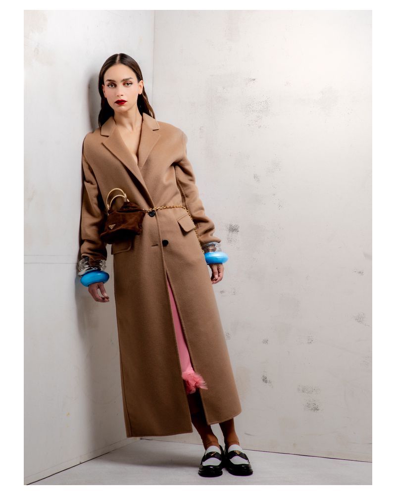 A woman in a tan coat leans against a corner, accessorized with blue bracelets and a mini bag, highlighted by studio lighting.