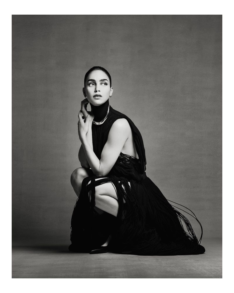 Elegant woman in a black fringed dress poses in a neutral studio. The portrait captures a modern editorial style, emphasizing fashion and portraiture.