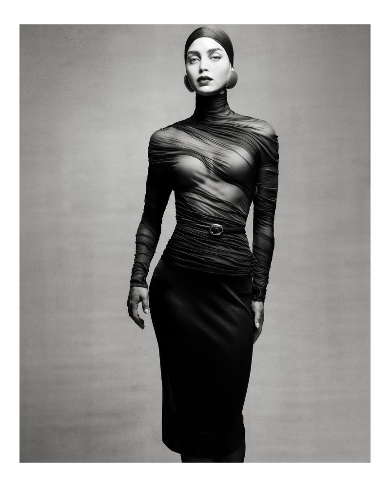 Monochrome portrait of a woman in a sheer top and skirt, accessorized with large earrings and a head covering against a neutral backdrop.