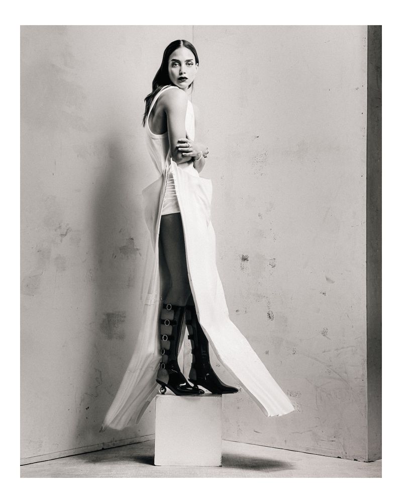 Monochrome fashion shot of a slender woman in a white dress with black boots posing on a white cube in a studio setting.
