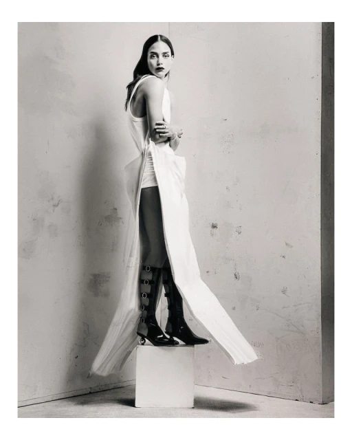 Monochrome fashion shot of a slender woman in a white dress with black boots posing on a white cube in a studio setting.