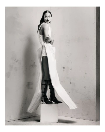 Monochrome fashion shot of a slender woman in a white dress with black boots posing on a white cube in a studio setting.
