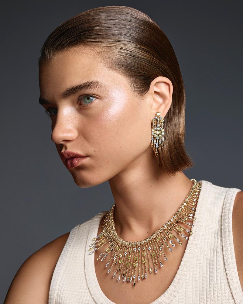 Close-up portrait of model Meghan Roche wearing elaborate diamond jewelry and a white ribbed top against a grey background.
