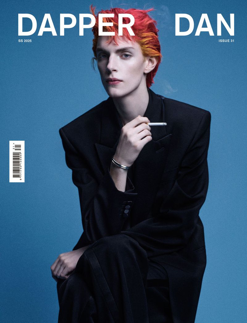 Magazine cover featuring a non-binary adult smoking, wearing a black suit against a light blue background with bright orange and yellow hair.