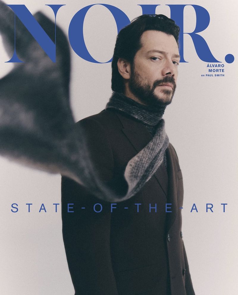 Magazine cover featuring a stylish adult male with a beard, wearing a dark coat and scarf, against a soft background with text elements.