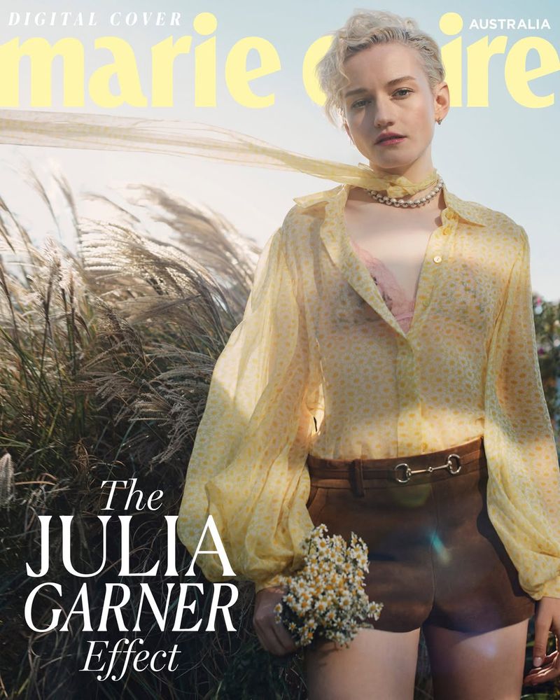 Julia Garner graces a magazine cover with a yellow chiffon blouse, brown shorts, and flowers, set against a soft outdoor backdrop.