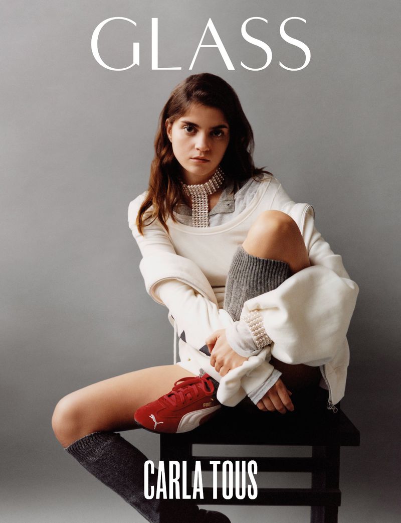 Fashion editorial portrait of Carla Tous sitting on a stool, wearing casual outfit with red Puma shoes, against a grey background.