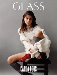 A seated woman wearing layered white clothing and red sneakers against a plain grey backdrop for Glass Magazine