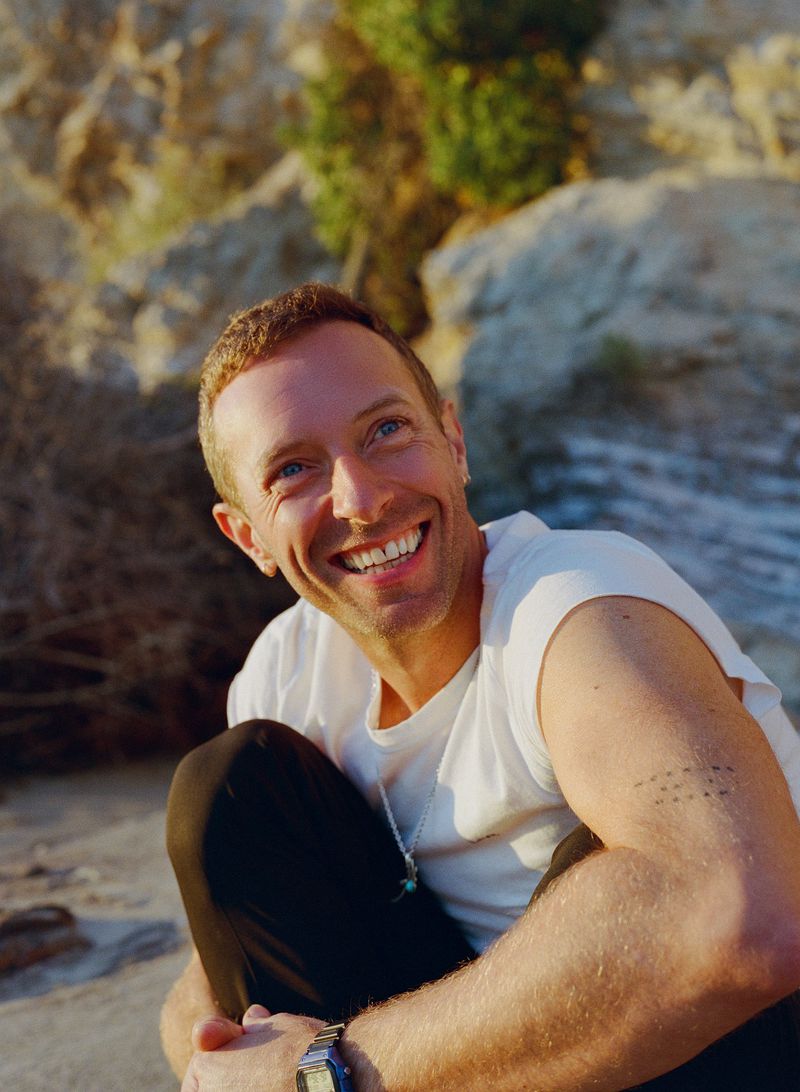 Portrait of a fair-skinned adult man with short hair smiling while sitting outdoors on a sandy surface with rocky formations.
