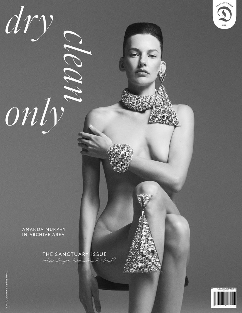 Black and white fashion magazine cover featuring a nude woman adorned with jeweled collar, bracelet, and posing on stool.