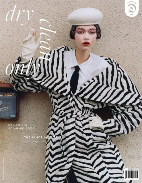 Fashion editorial: A model poses with a cigarette in a black and white patterned coat, white collar shirt, and white beret, against a neutral background.