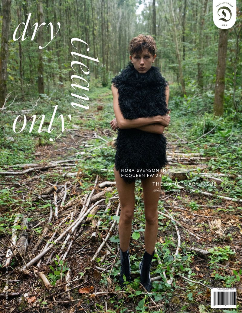 Fashion editorial image featuring a model in a black, textured outfit against a backdrop of dense green woods and natural ground cover.