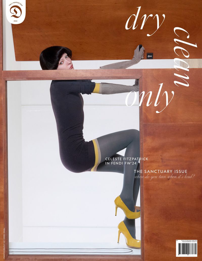 Fashion magazine cover featuring a woman in grey and yellow clothing, perched in a wooden frame, with a white background and bold text.