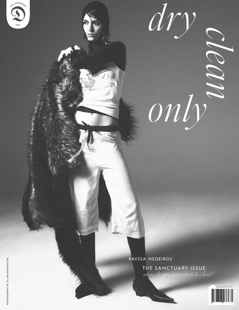 Fashion shot of a female model in a light-colored outfit and black boots holding a long fur coat against a gray background.