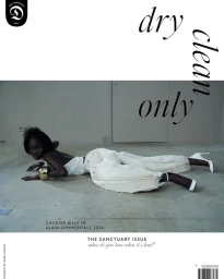 Model lying on concrete floor wearing white top, pleated pants, and white heels, holding a large gold bangle