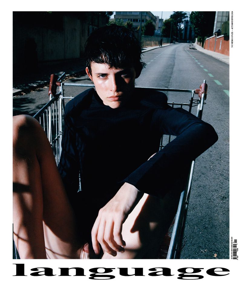 A young adult woman is sitting in a shopping cart in the middle of an empty road wearing a dark top, captured in natural lighting.