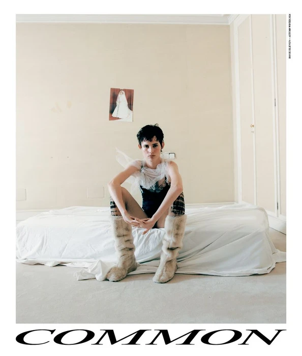 Model with short dark hair wearing a black bodysuit and furry boots seated on a white bed in a domestic bedroom