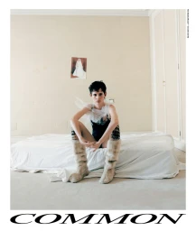 Model with short dark hair wearing a black bodysuit and furry boots seated on a white bed in a domestic bedroom