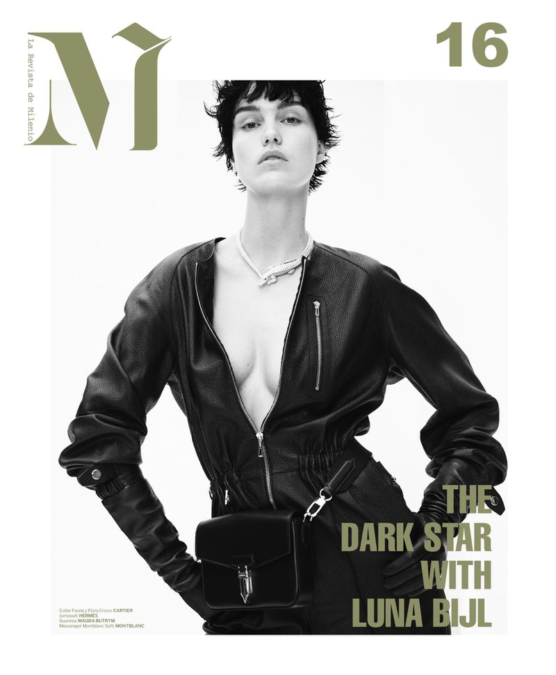 Fashion editorial shot of a young woman in a black leather jumpsuit, gloves, and bag, accessorized with a silver necklace against white.