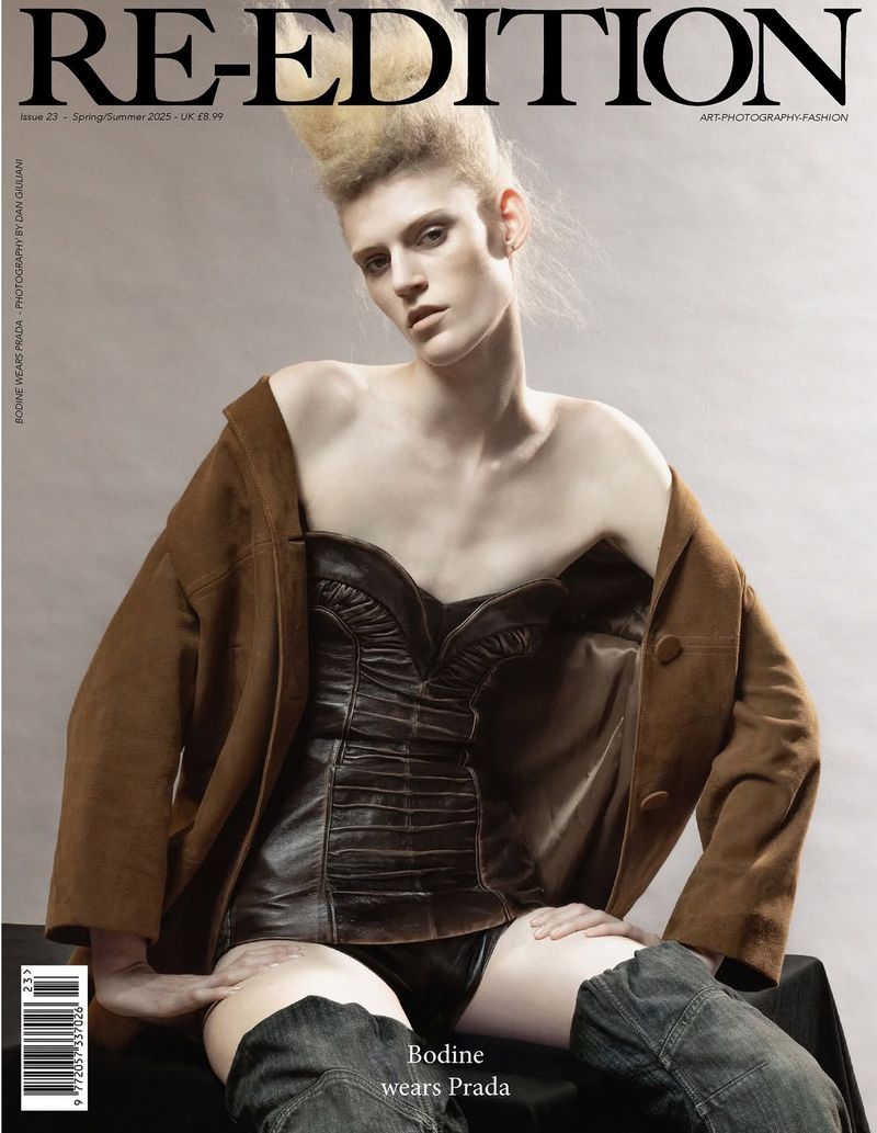 Magazine cover featuring a posed person with light skin, wearing a brown jacket and black corset, seated against a neutral background.
