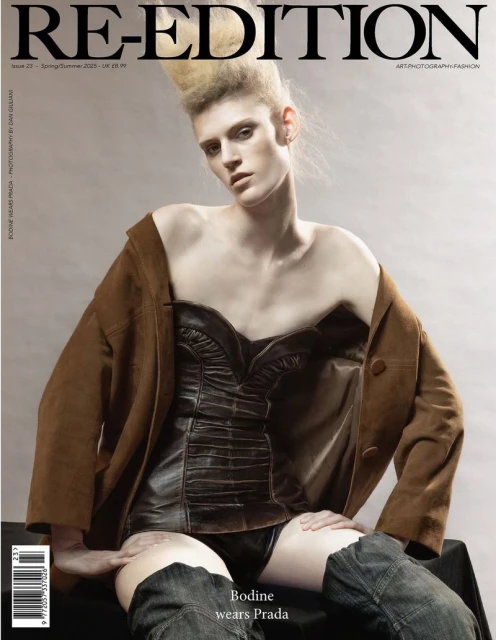 Magazine cover featuring a posed person with light skin, wearing a brown jacket and black corset, seated against a neutral background.