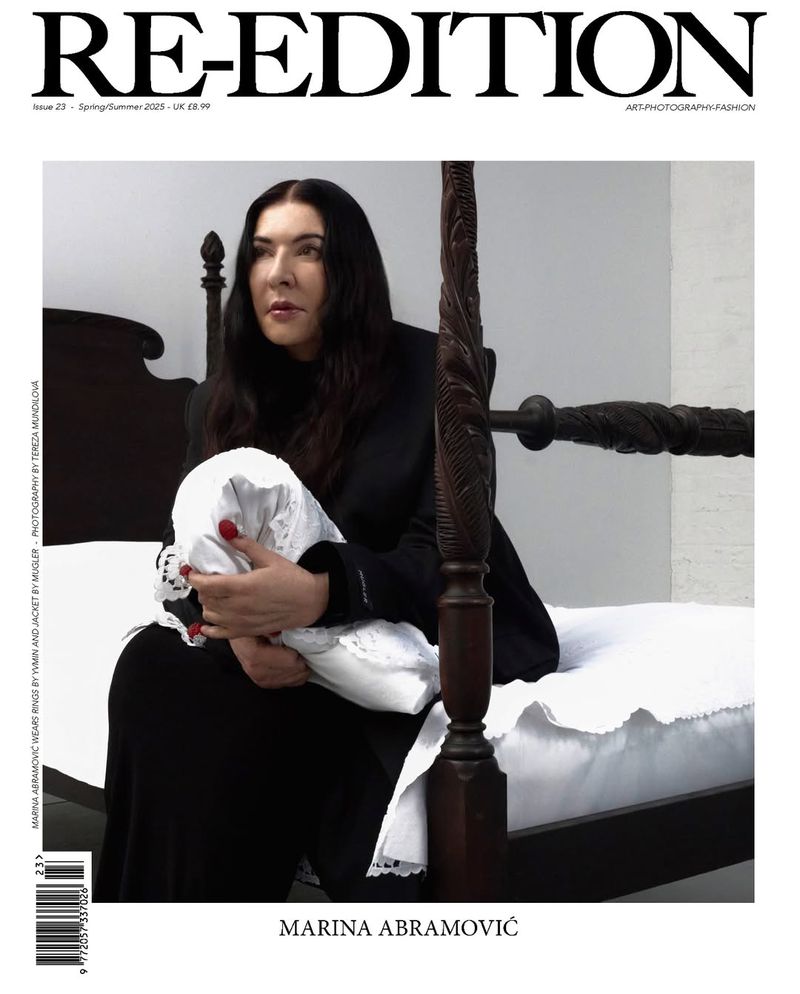 Magazine cover featuring Marina Abramović seated on a dark wood bed holding a white folded fabric in a minimalist setting.