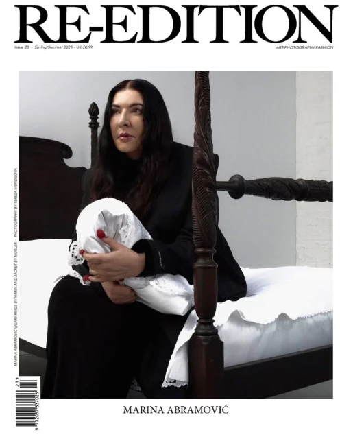Magazine cover featuring Marina Abramović seated on a dark wood bed holding a white folded fabric in a minimalist setting.