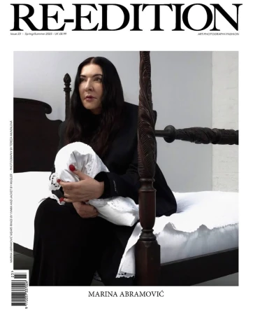 Magazine cover featuring Marina Abramović seated on a dark wood bed holding a white folded fabric in a minimalist setting.