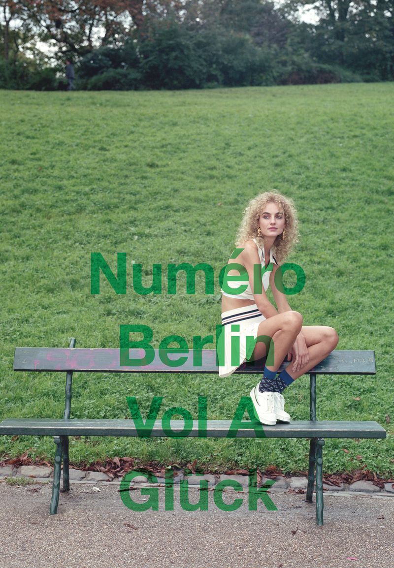A woman with curly blonde hair sitting on a park bench in a white athletic outfit and sneakers, green grass and trees in background.