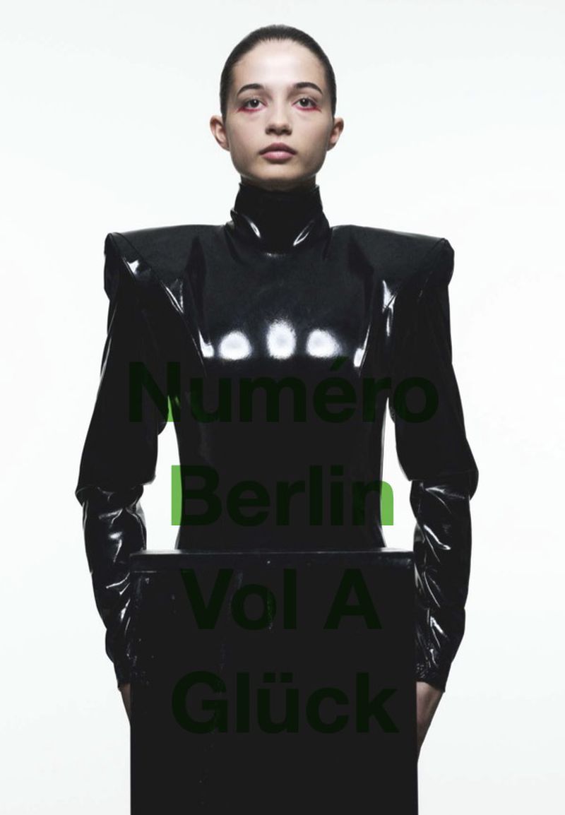 Fashion editorial portrait of a woman in a black vinyl dress against a bright white backdrop, featuring magazine title text overlay.