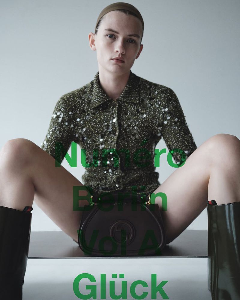 A model sits indoors on a platform wearing a green sequined jacket and dark boots with a dark handbag with overlaid text in front of a white wall.