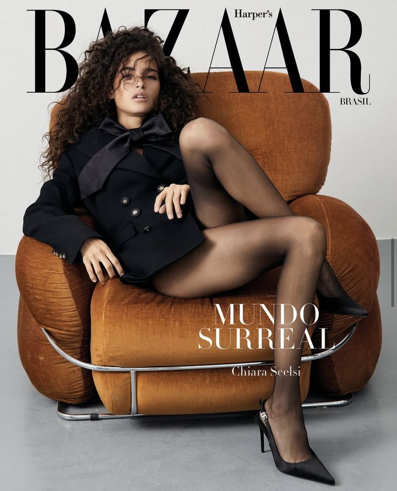 Magazine cover featuring a woman with curly hair posing in an armchair. She is wearing a black blazer, black tights, and high heels.