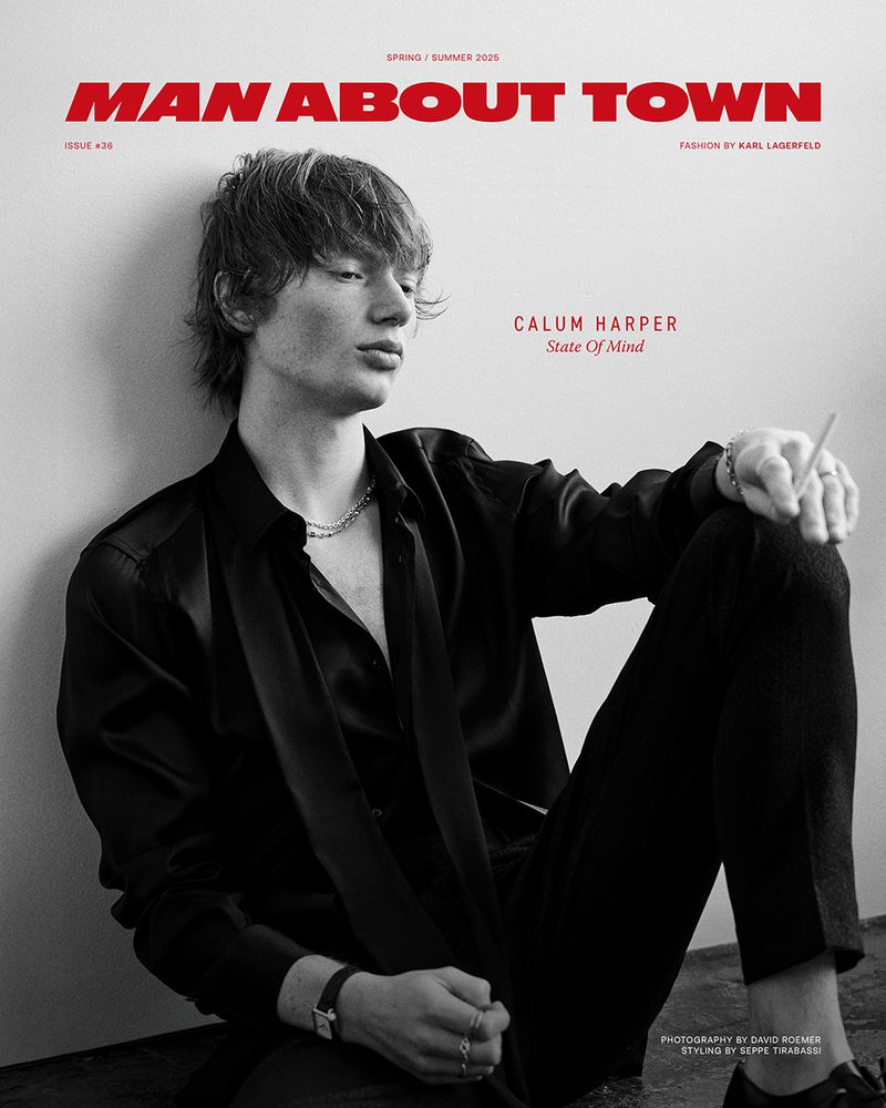Black-and-white fashion editorial shot of a young man with dark hair in a black shirt and pants, posed against a light wall, holding cigarette.