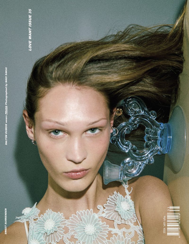 Close-up of a woman with long brown hair, light skin, and light blue floral dress. She has blue glass objects in her hair.